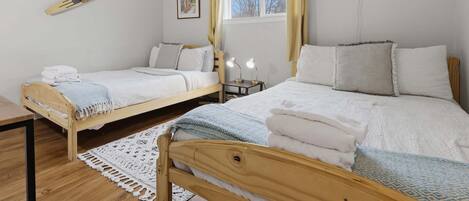 Un dormitorio con dos camas, piso de madera, ventana con cortinas, mesita de noche con lámpara y aire acondicionado instalado en la pared.