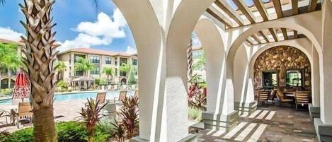 A covered outdoor area with a pool, palm trees, and a patio with chairs and tables.