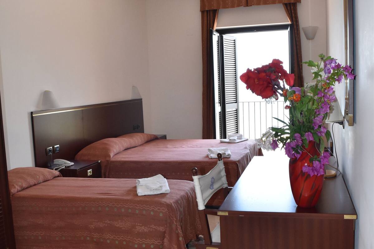 Habitación de hotel con dos camas, un escritorio, un florero con flores y una ventana con cortinas.