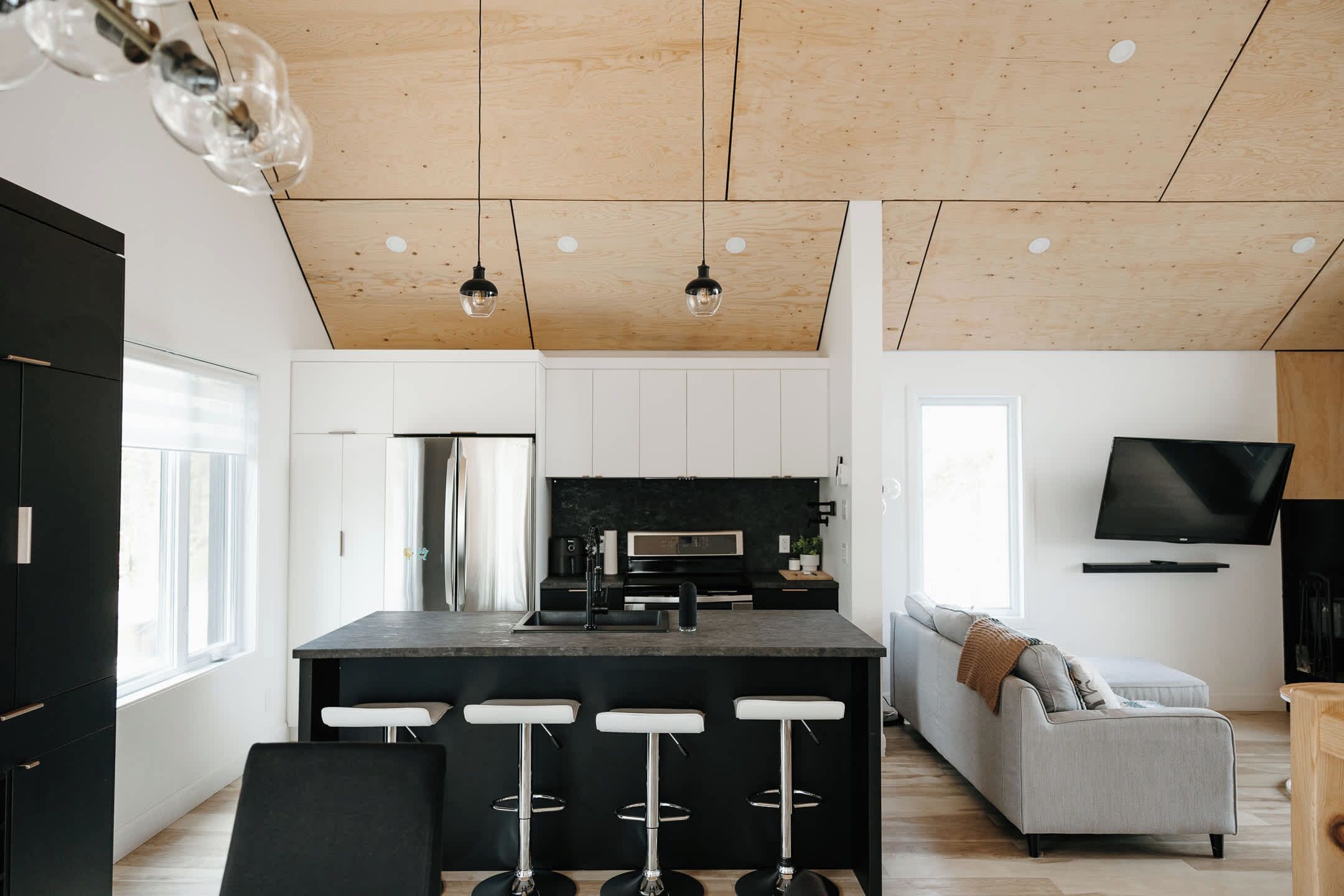 Una cocina moderna con una isla central, taburetes de bar, un sofá gris, un televisor de pantalla plana y un techo de madera.