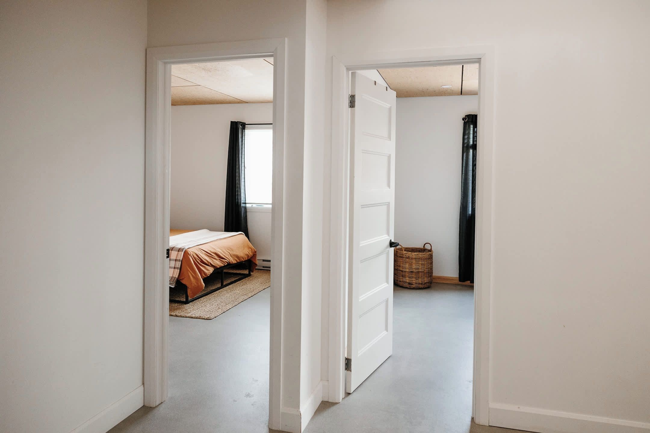 Un couloir mène à une chambre où se trouvent un lit, une fenêtre avec des rideaux et un panier en osier posé sur le sol.