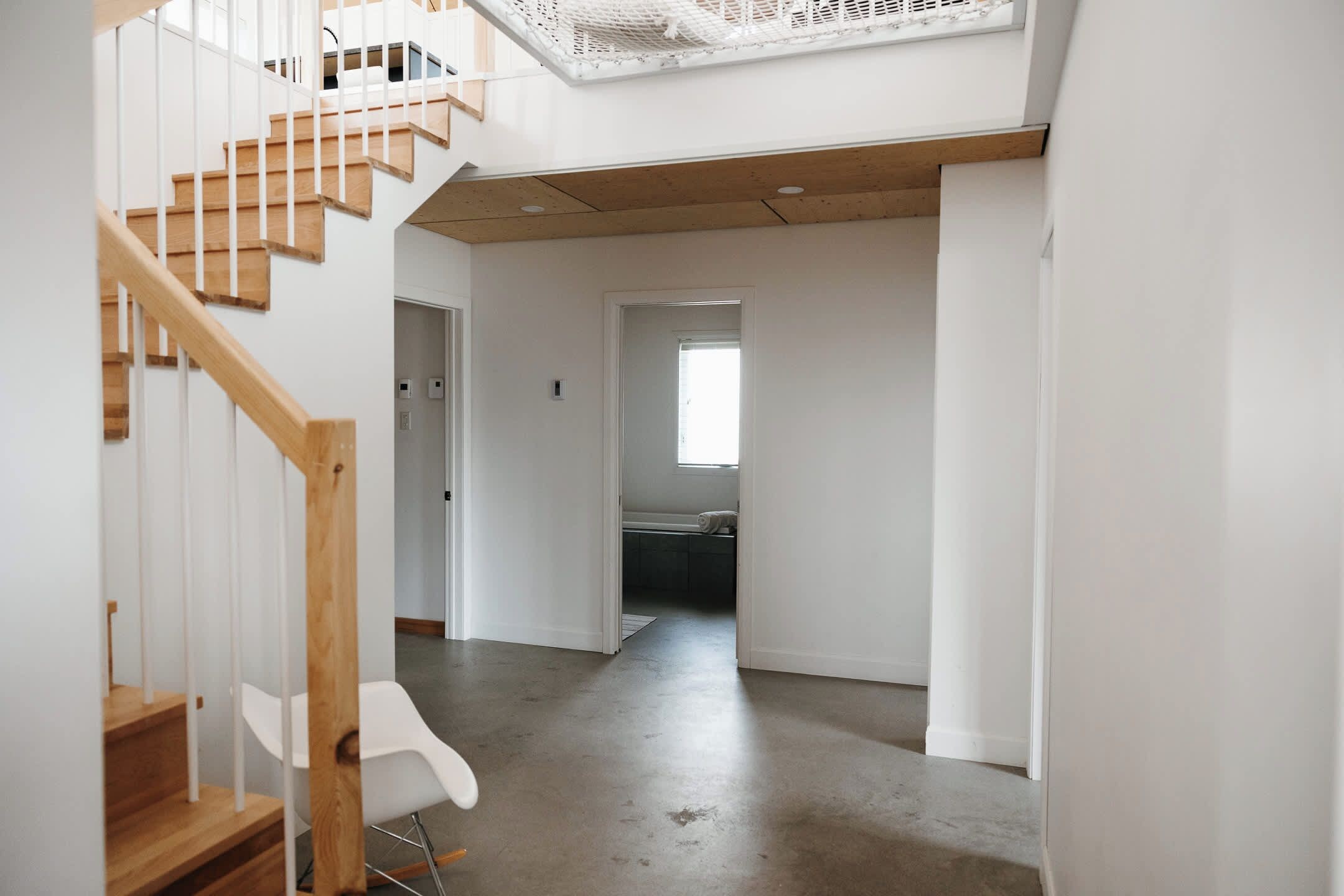 A modern interior with a wooden staircase, a white chair, and a view into a kitchen area.