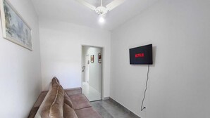 A room with a brown sofa, a ceiling fan, a TV mounted on the wall, and a door leading to another room. - Hostel Vila Univesitária (Bauru)