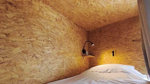A small, cozy room with wooden walls, a single bed with white bedding, and a wall-mounted light fixture. - Hostel Vila Univesitária (Bauru)