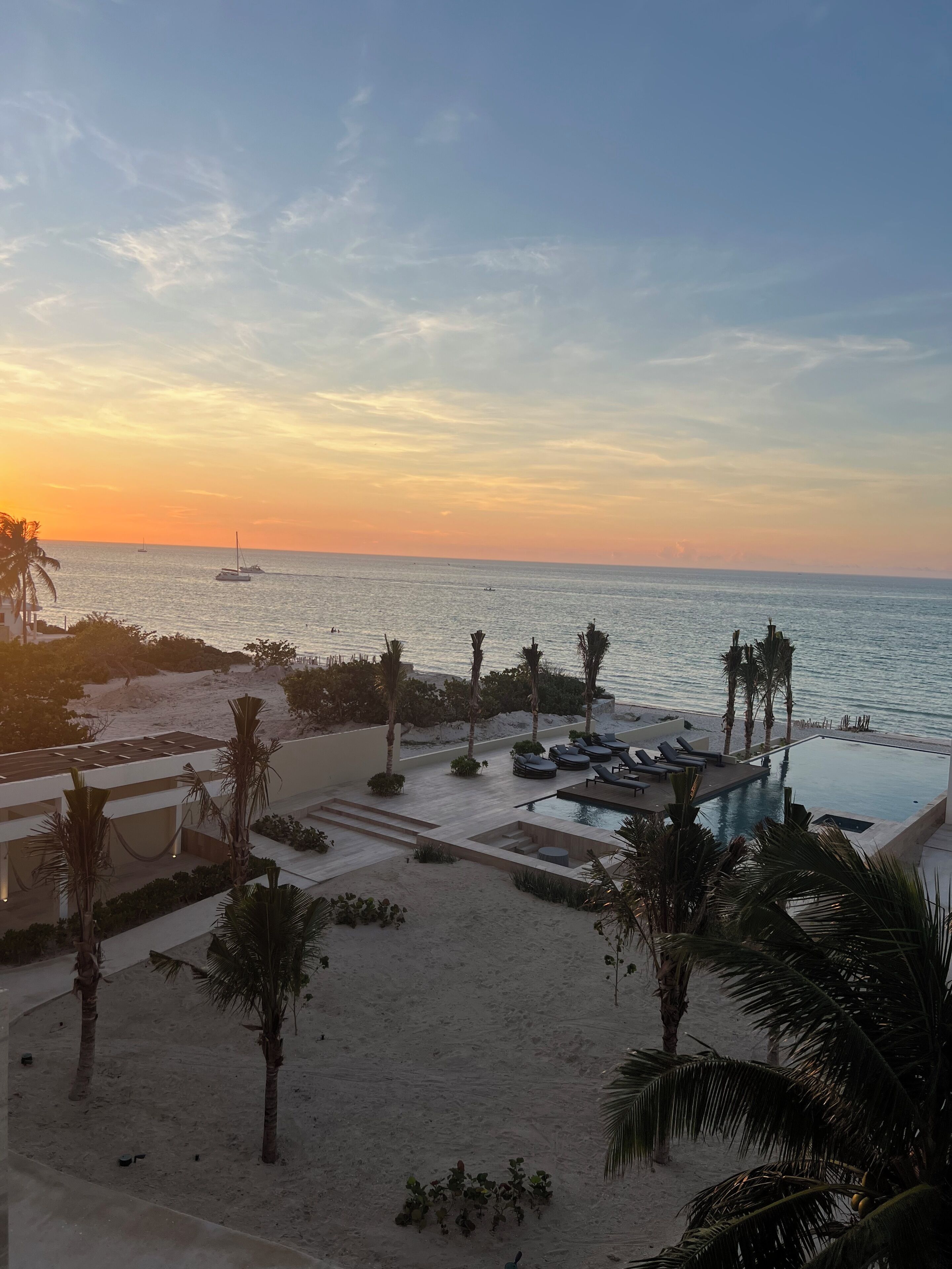 A resort with a pool, lounge chairs, palm trees, and a view of the ocean at sunset.