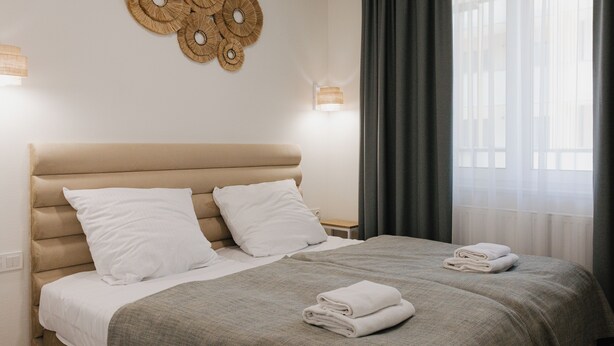 A neatly made bed with white pillows and a grey blanket, a wall-mounted light fixture, and a window with curtains. - Apartamenty Sun&Snow Aquamarina Dąbki (Dąbki)