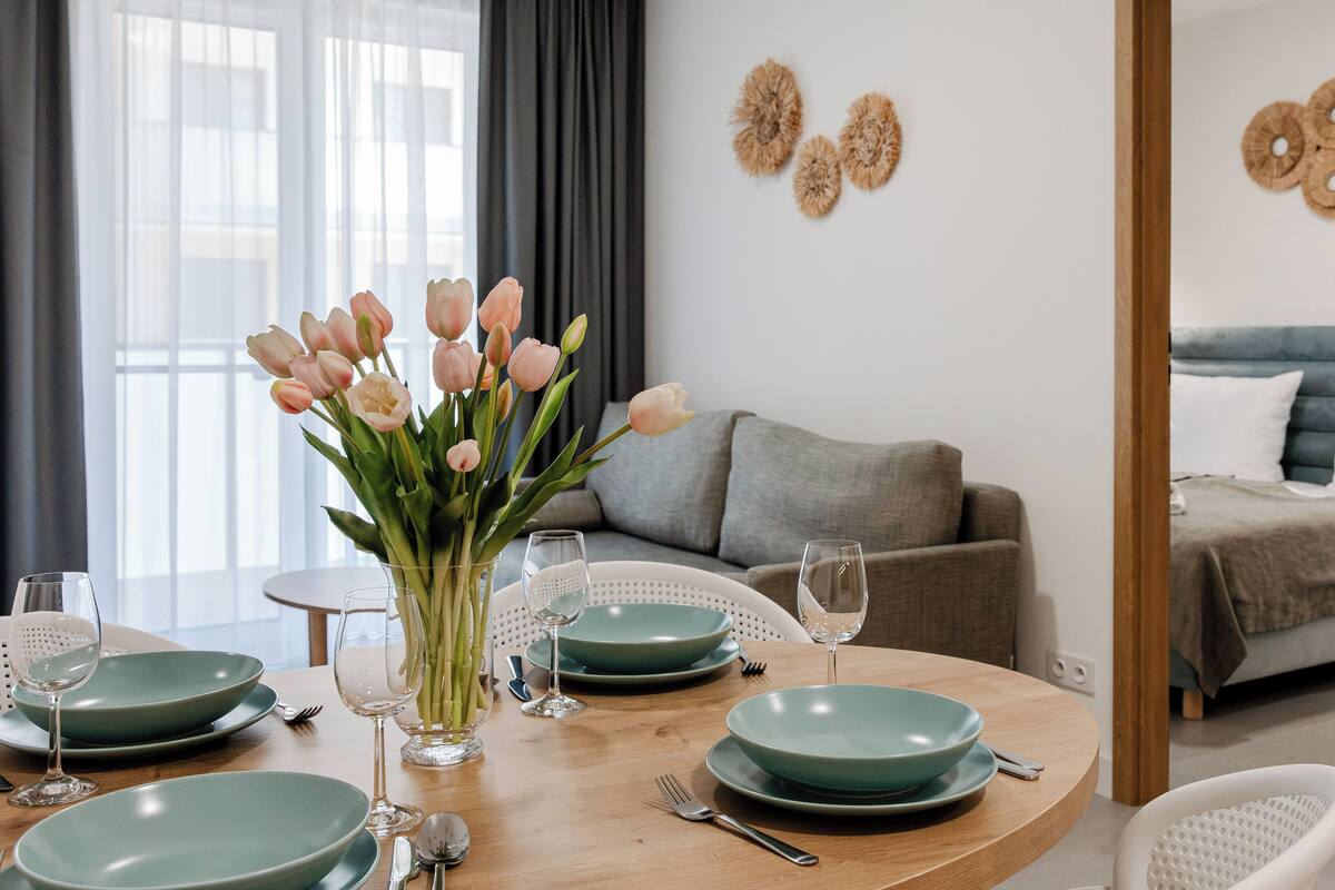 A dining area with a round table set with plates, glasses, and cutlery. There is a vase with flowers on the table.