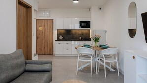 A compact living space with a kitchenette, a dining area with white chairs, a grey sofa, and a wooden door. - Apartamenty Sun&Snow Aquamarina Dąbki (Dąbki)