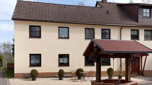 A two-story building with a tiled roof, multiple windows, a small outdoor seating area with chairs and tables, and a wooden structure with a sloped roof. - Apartment “Linke Hof” with Wi-Fi (Bobritzsch-Hilbersdorf)