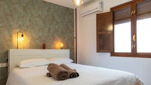 Una cama bien tendida con ropa blanca y dos toallas enrolladas, un aplique de pared y una ventana con contraventanas de madera.
