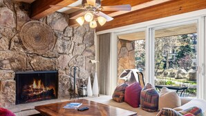 A stone fireplace, a wooden ceiling fan, a stone wall, a sofa with colorful cushions, and a view of a garden through a large window. - Cozy Mountain Getaway by InvitedHome | Walk to Ski, Hot Tub, Village Center (Vail)