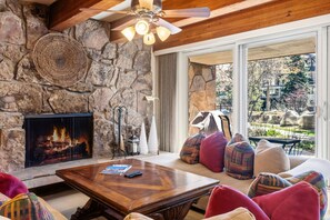 A stone fireplace, a wooden ceiling fan, a stone wall, a sofa with colorful cushions, and a view of a garden through a large window. - Cozy Mountain Getaway by InvitedHome | Hot Tub, Pool, Village Center (Vail)