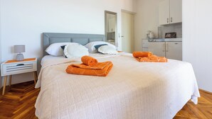 Una cama bien hecha con una manta blanca y naranja, una mesita de noche con una lámpara, y una zona de cocina con armarios.