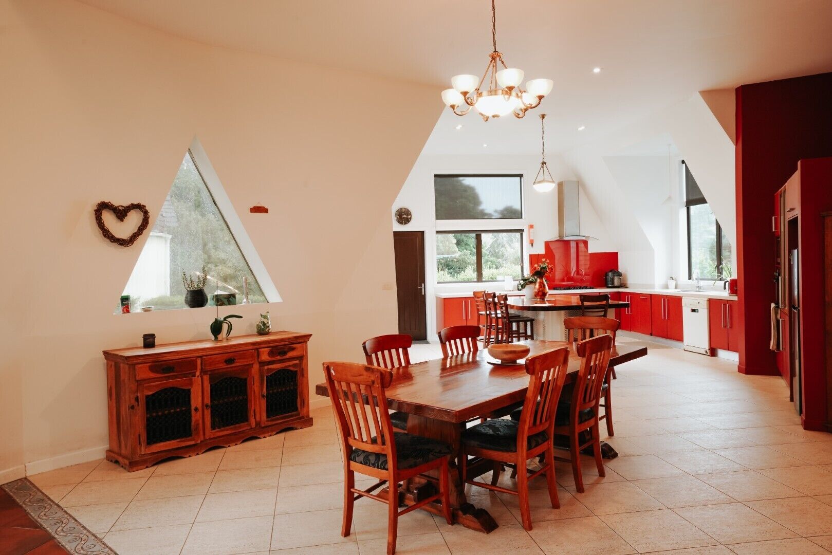Un coin repas avec une table et des chaises en bois, un lustre et une cuisine dotée de meubles rouges et d’une fenêtre.