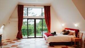 Un dormitorio con cama, una silla, una mesita de noche, un ventanal con cortinas y vistas al exterior.