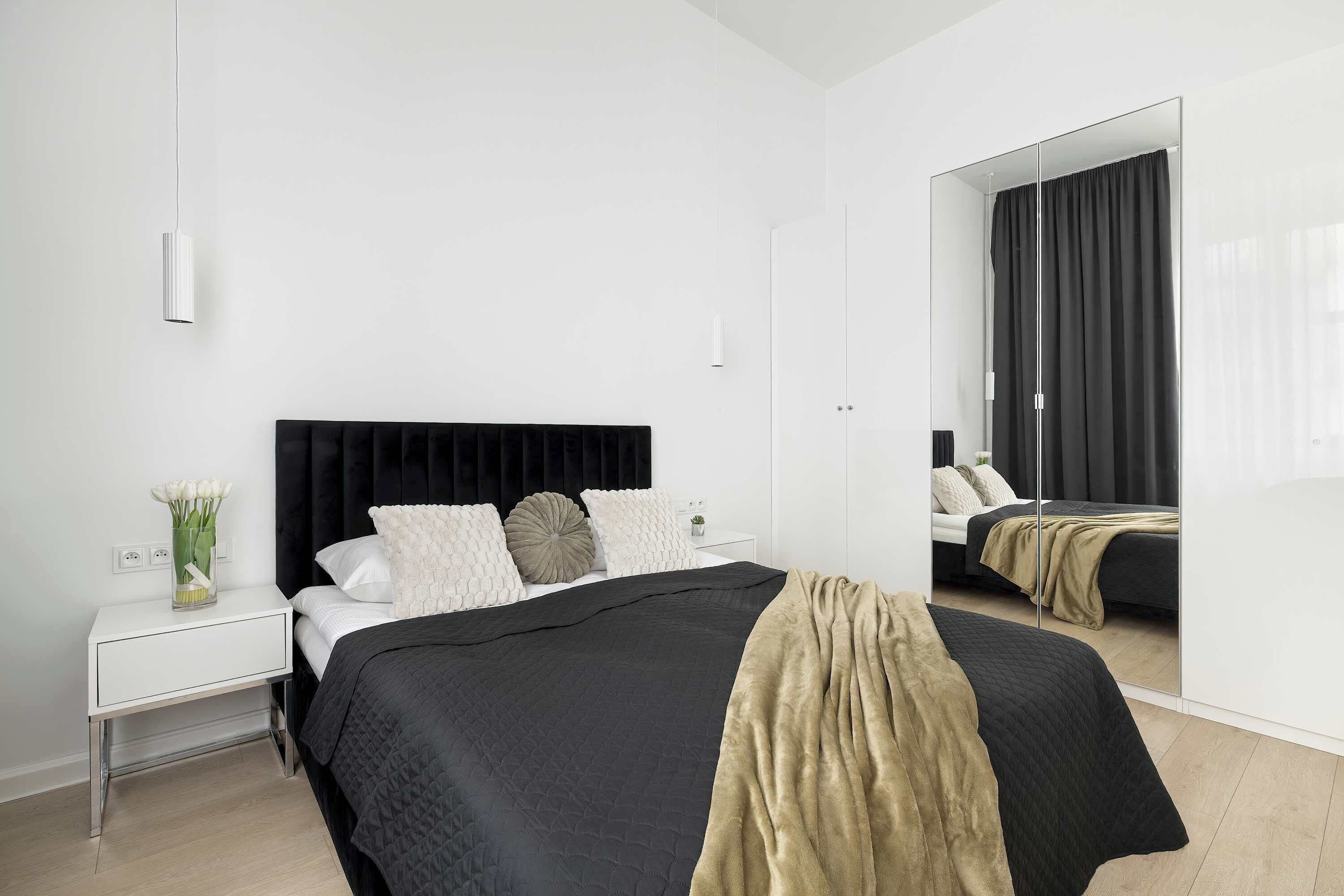 A bright and minimalist bedroom features a large bed with a dark duvet and a decorative gold throw. A tall, dark mirror stands in the corner, reflecting the clean, white walls of the room.