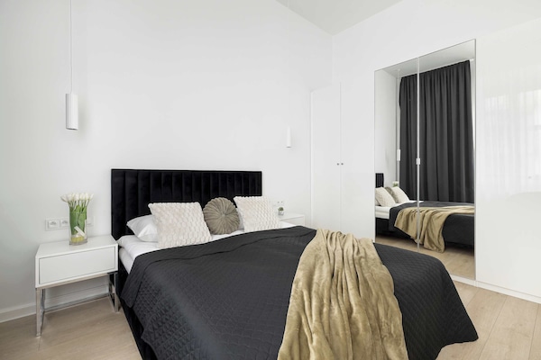 A bright and minimalist bedroom features a large bed with a dark duvet and a decorative gold throw. A tall, dark mirror stands in the corner, reflecting the clean, white walls of the room.