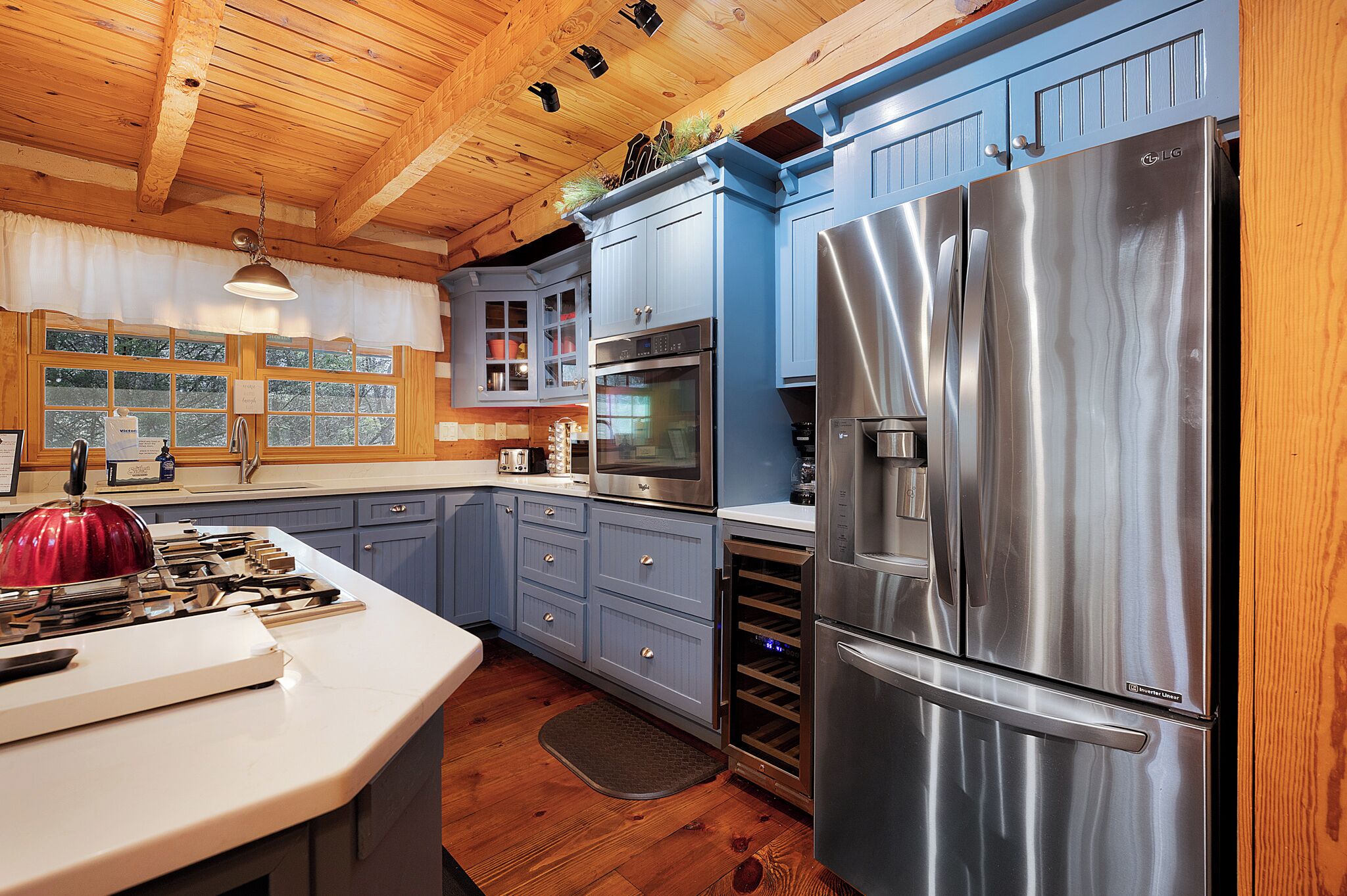 A kitchen with wooden ceiling beams, a stainless steel refrigerator, a built-in oven, and a large island with a red kettle.