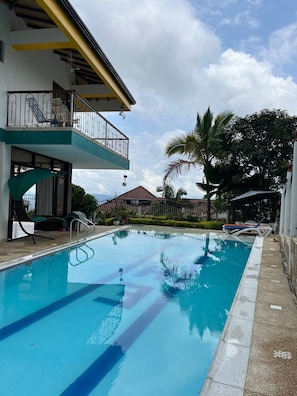 Una piscina con sillas de descanso, un espacio de estar cubierto y un edificio con balcón.