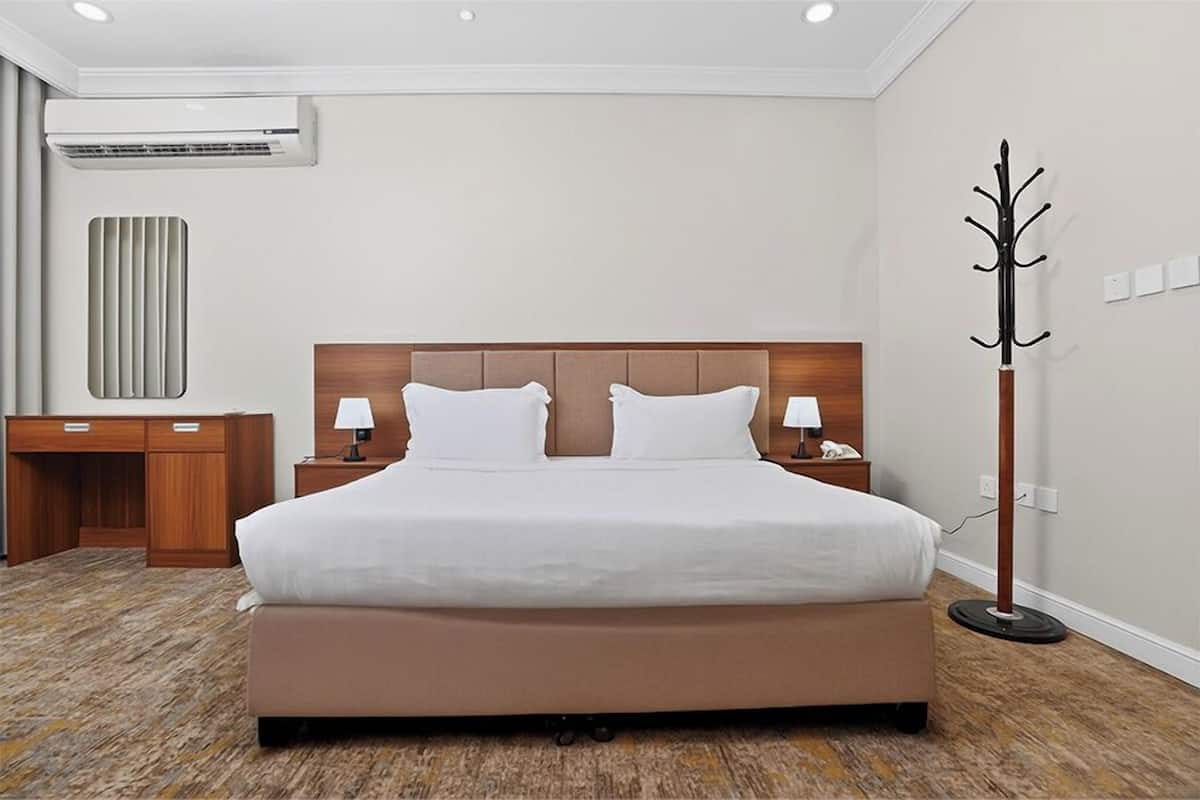 Ein Hotelzimmer mit einem großen Bett, Nachttischen, einem Schreibtisch, einer Klimaanlage, einem Kleiderständer und einer Wandleuchte.