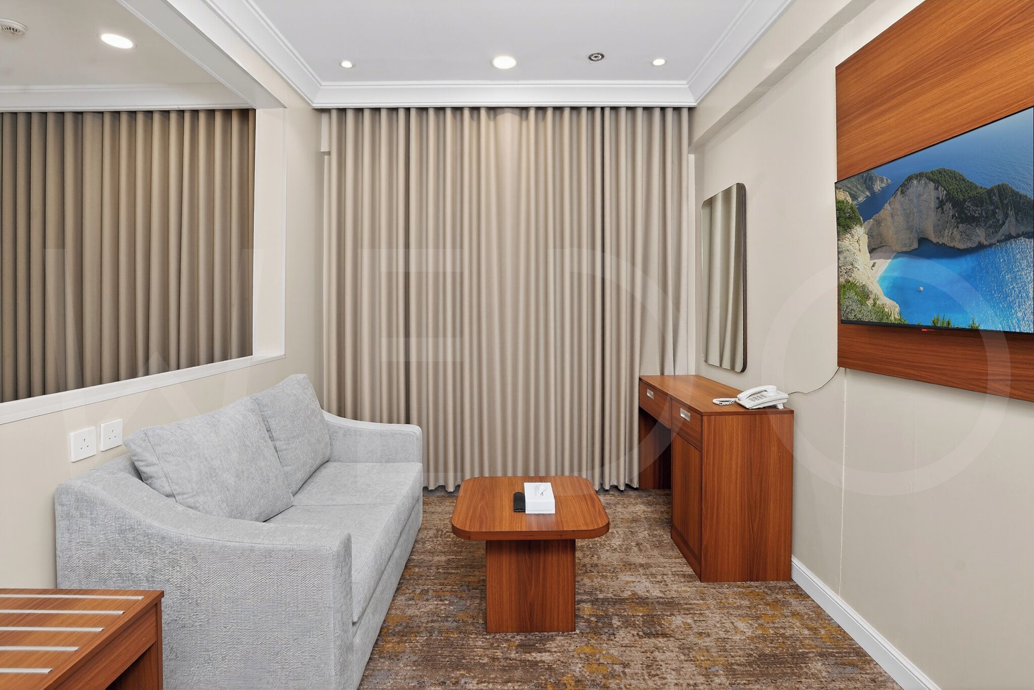 Une chambre d’hôtel avec un canapé gris, une table basse en bois, un bureau avec un téléphone et un grand tableau représentant un paysage côtier.