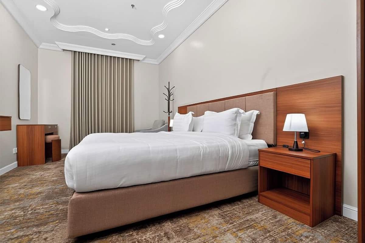 Ein Hotelzimmer mit einem großen Bett, einem Nachttisch aus Holz mit einer Lampe, einem Kleiderhaken und einem Fenster mit Vorhängen.