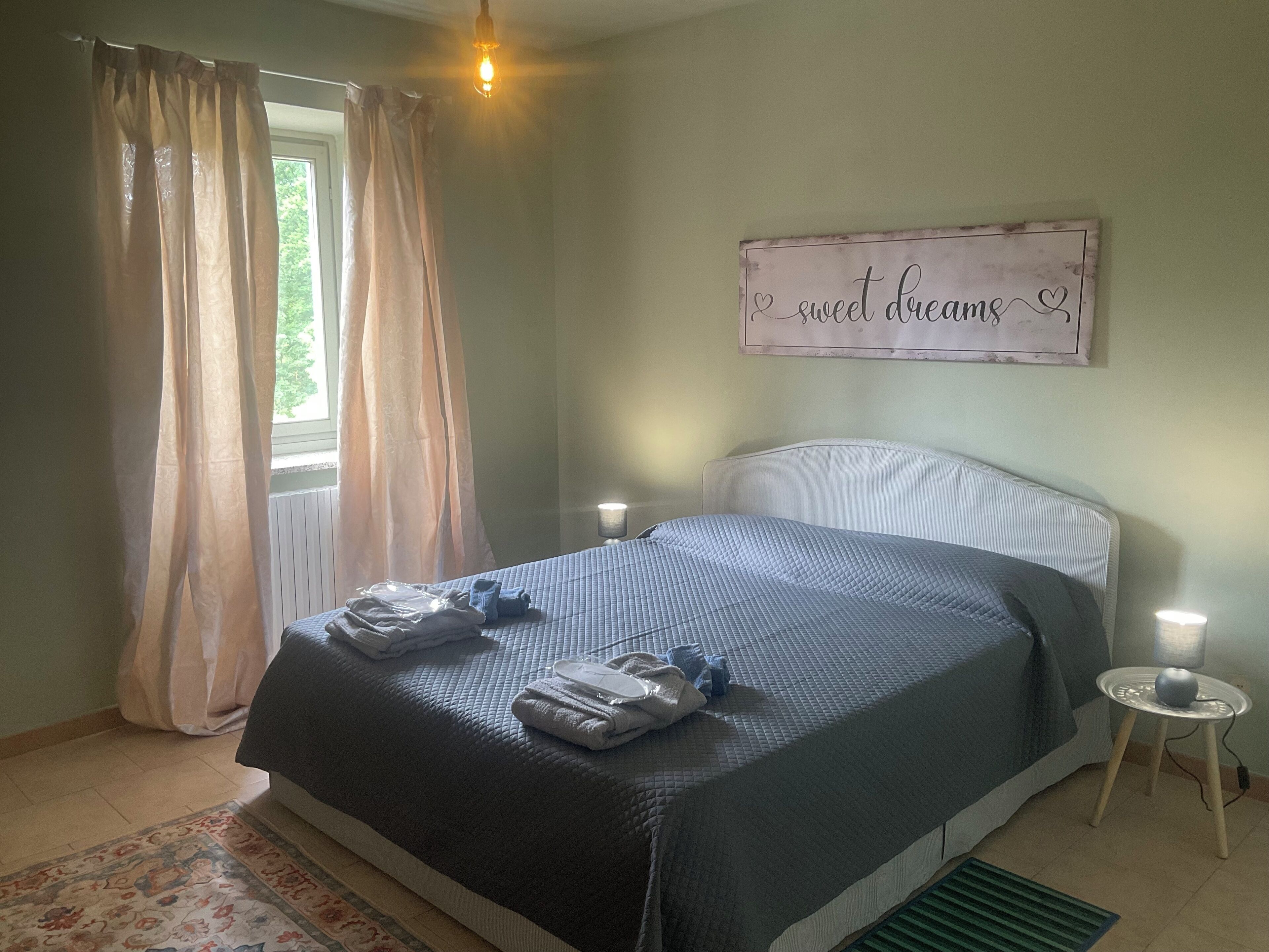 Ein Schlafzimmer mit einem Bett, zwei Nachttischlampen, einem fensterbelaubten Fenster, einem Wandschild und einem kleinen Tisch mit einer Lampe.