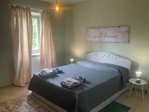 Une chambre Ă coucher avec un lit, deux lampes de chevet, une fenĂȘtre avec des rideaux, un tableau mural et une petite table avec une lampe.