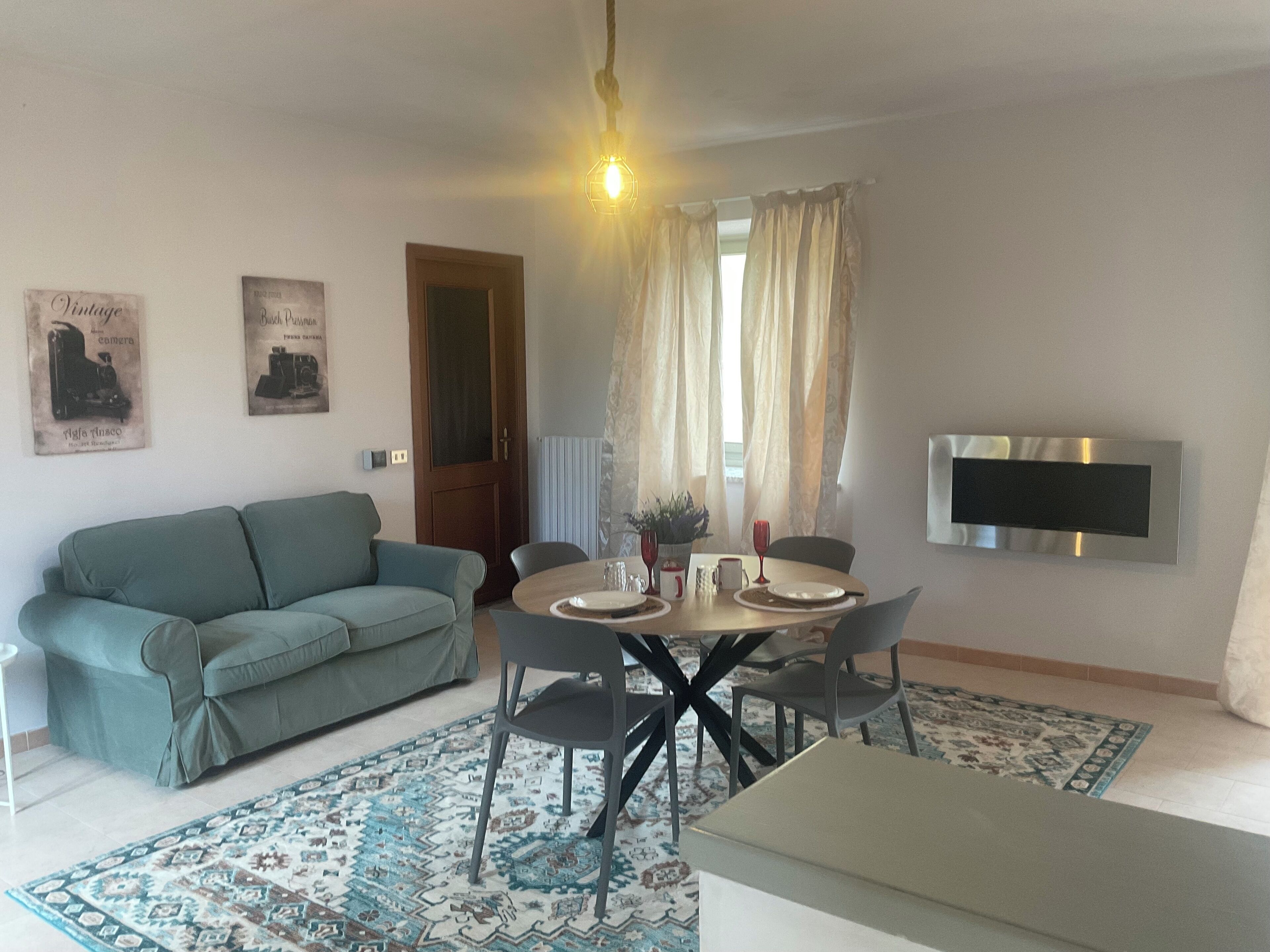 A living room with a sofa, a dining table with chairs, a TV mounted on the wall, and a patterned rug.