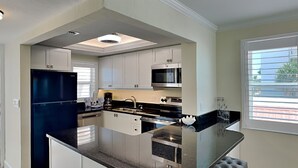 Una cocina moderna con armarios blancos, encimera negra, microondas integrado y una ventana con persianas.