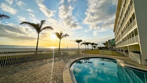 Zona de piscina con vista al océano y palmeras, un edificio con balcones y cielo despejado.