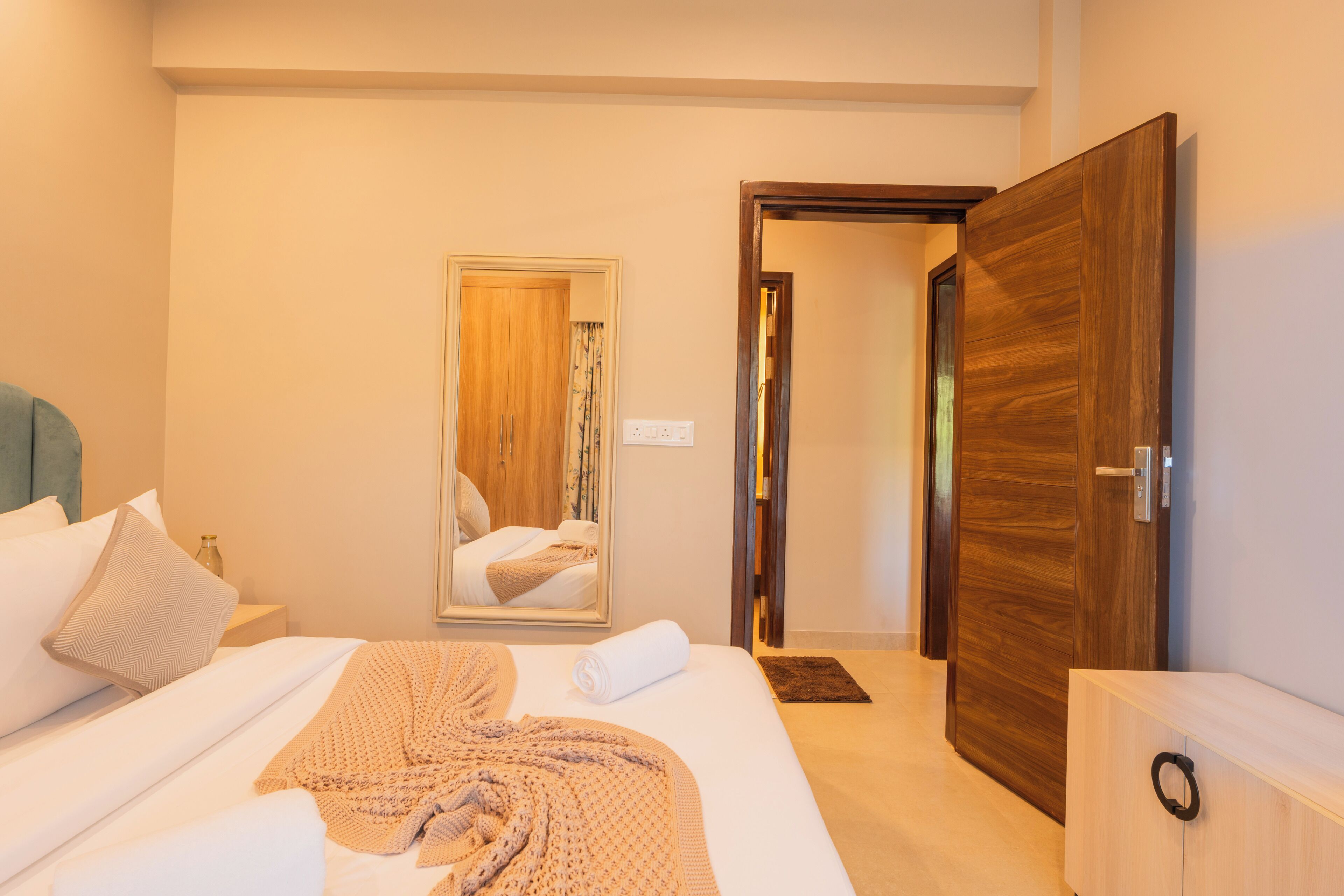 A hotel room with a bed, a wooden wardrobe, a mirror, and a door leading to another room.