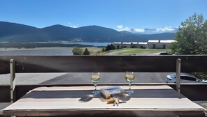 Una mesa con dos copas de vino, una botella y un recipiente pequeño en un balcón con vistas a un lago y montañas.