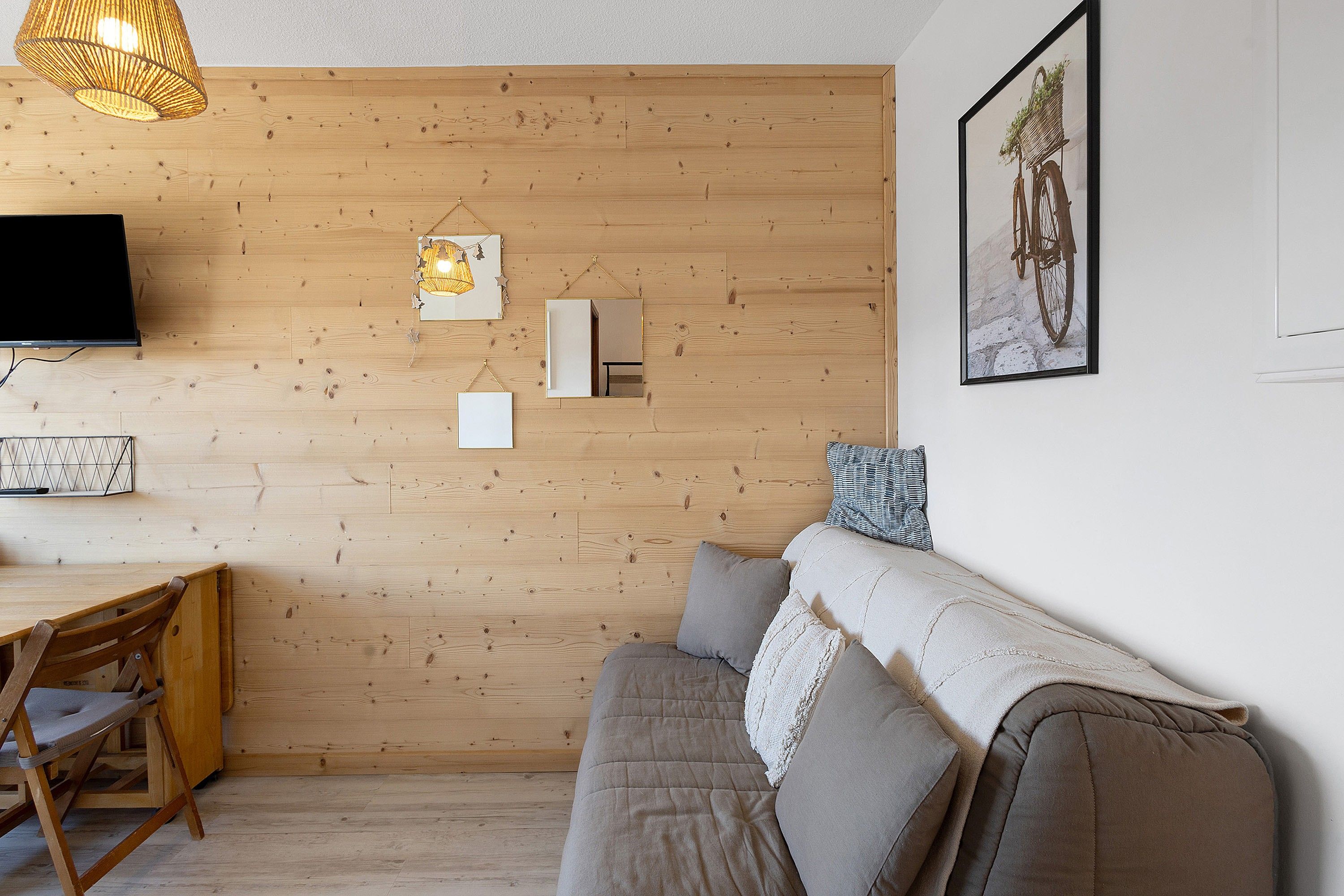 Una sala de estar moderna con una pared de madera, un televisor de pantalla plana, una zona de comedor con mesa y sillas de madera, y un sofá con cojines.