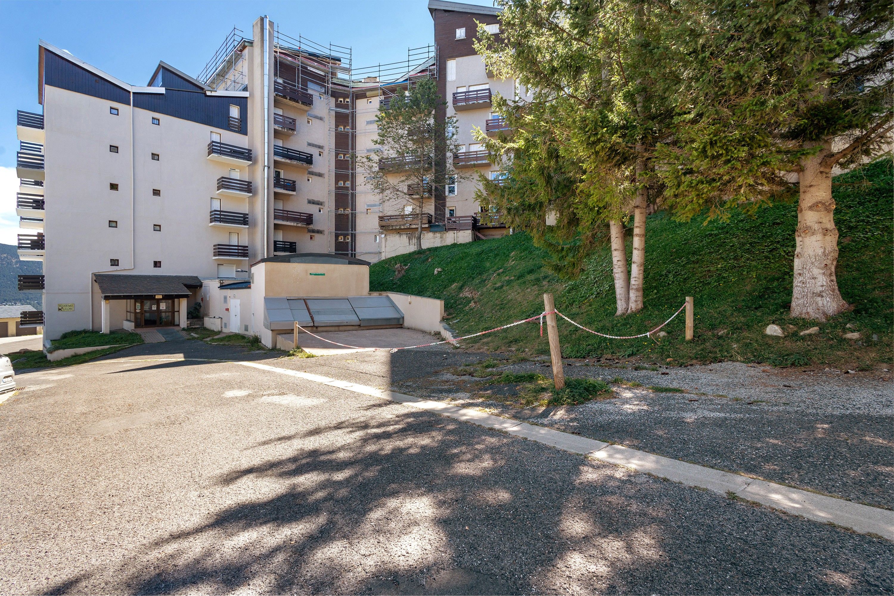 Un edificio residencial de varios pisos con entrada protegida, un pequeño estacionamiento y una colina cubierta de césped y árboles.