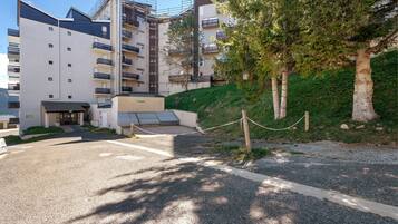 Un edificio residencial de varios pisos con entrada protegida, un pequeño estacionamiento y una colina cubierta de césped y árboles.