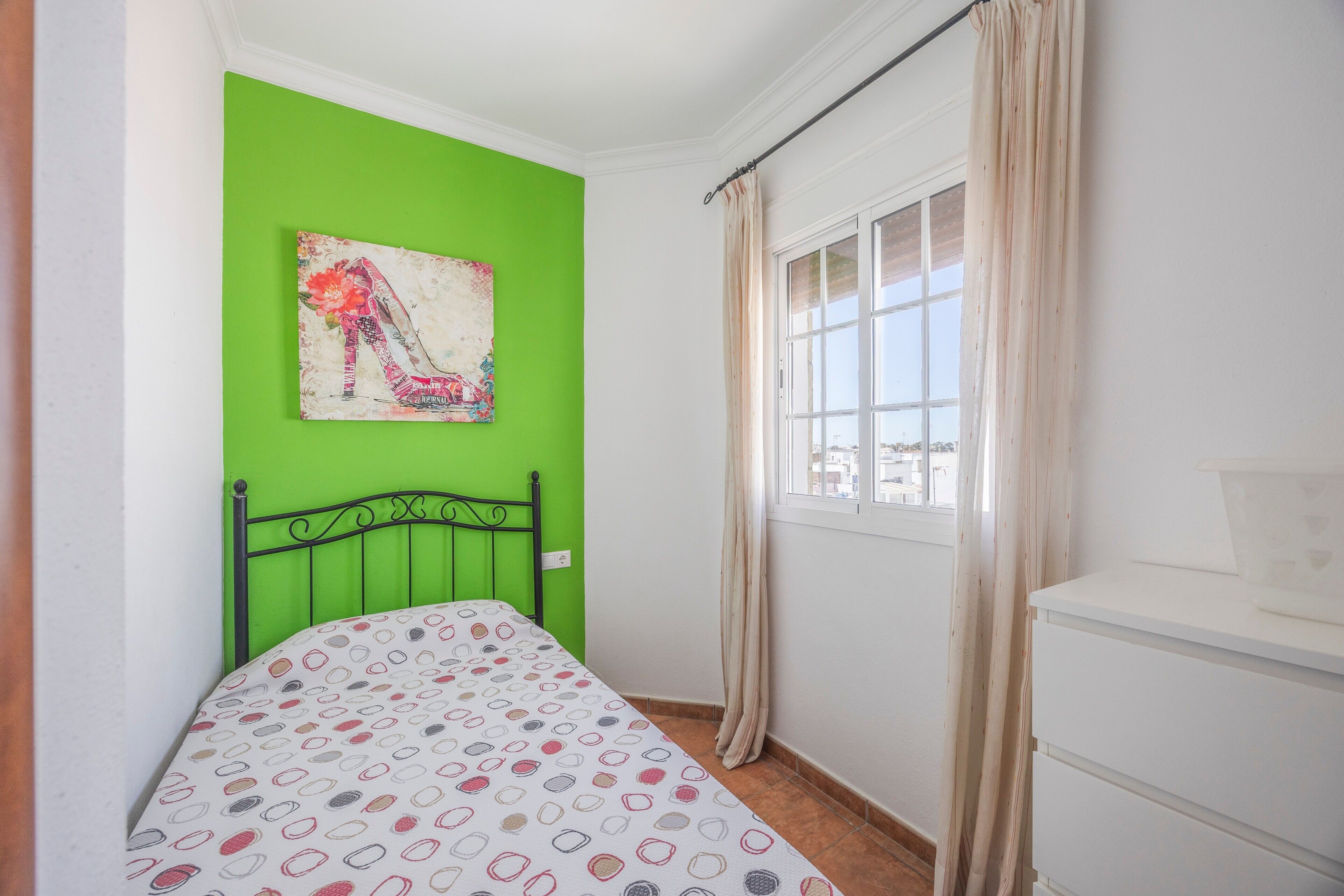 A room with a green wall, a bed with a patterned bedspread, a white dresser, and a window with curtains.