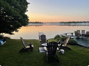 Ein Sitzbereich im Freien am Seeufer mit Adirondack-Stühlen und einem Tisch, umgeben von Gras und einem Baum.