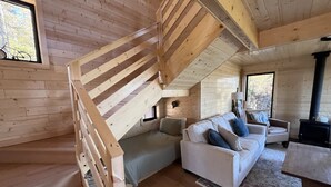 Ein Interieur einer Hütte mit Treppe, einer Sofagarnitur, einem Kamin und einem Fenster, vor dem ein Fahrrad steht.