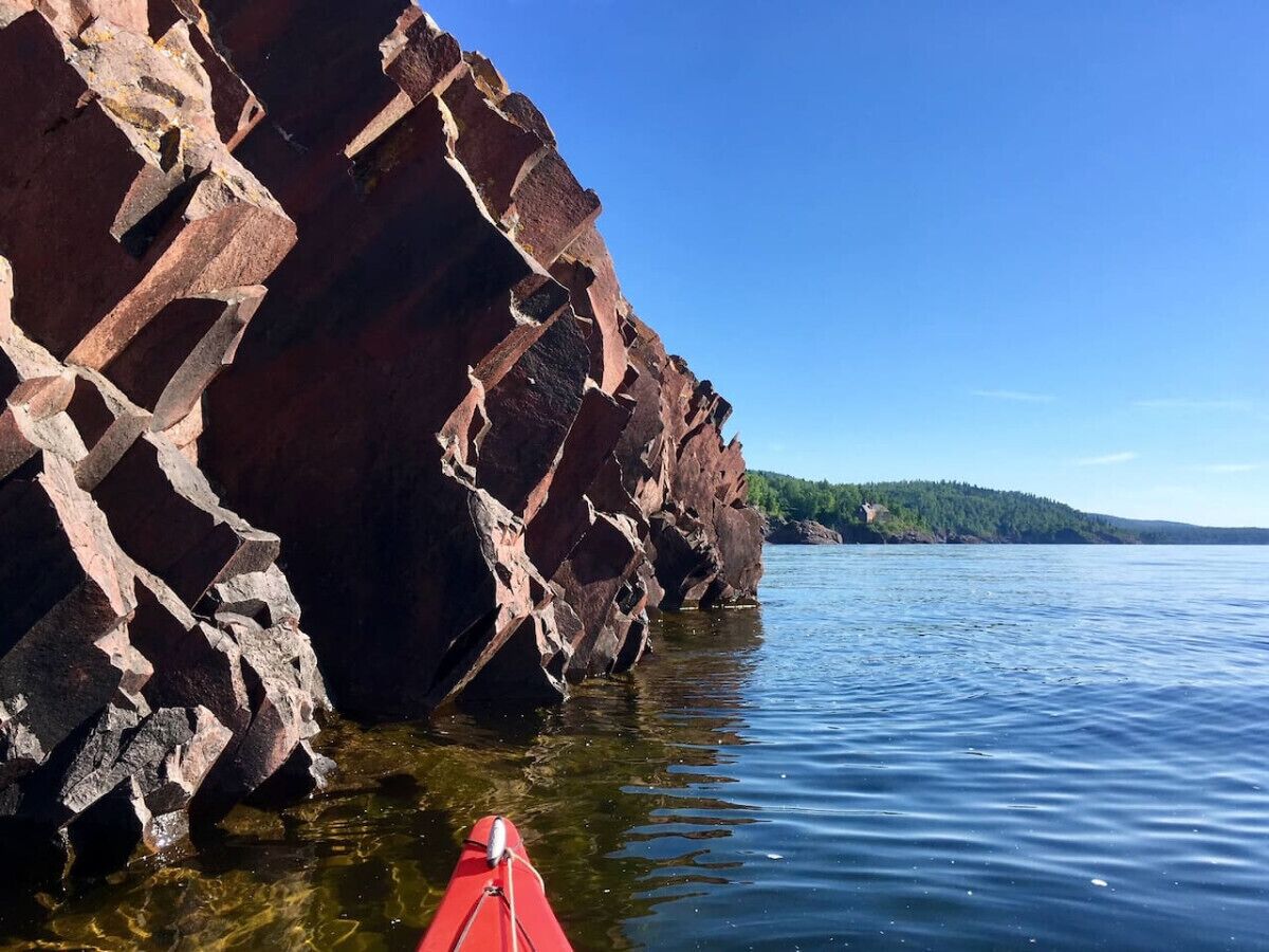 A kayaker in a red kayak near a rocky shoreline with clear water and a distant forested island.
