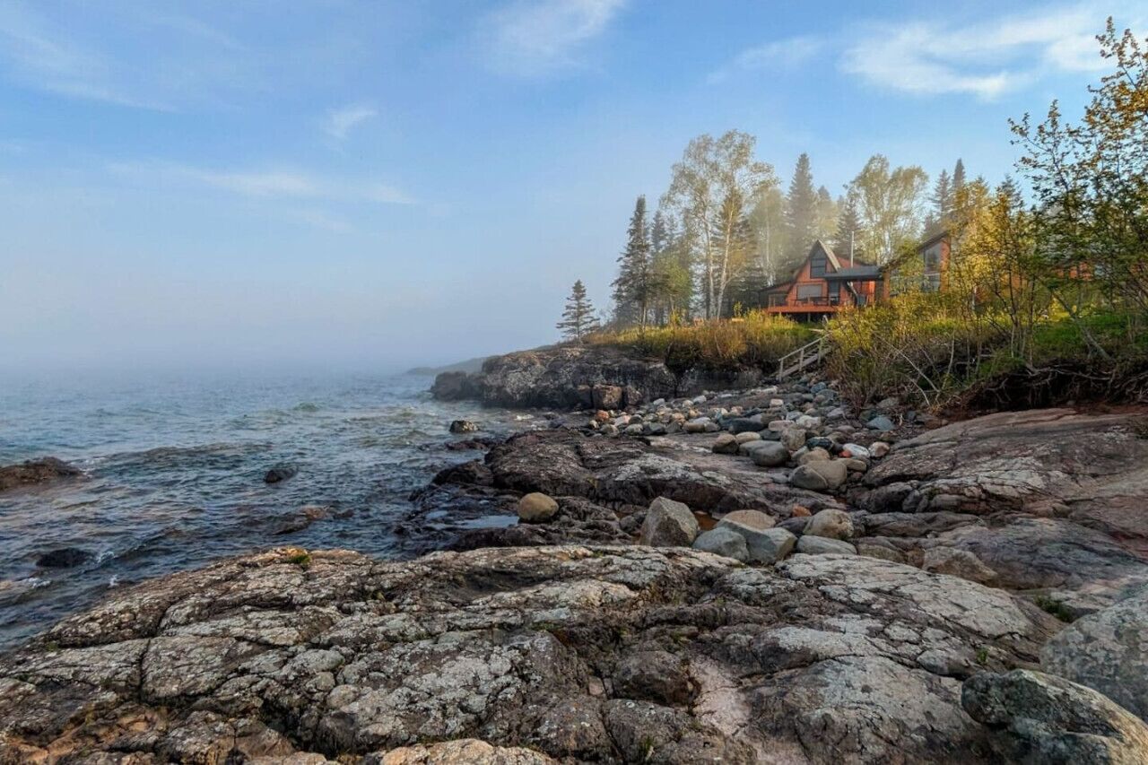A rocky shoreline with a cabin and trees in the background.