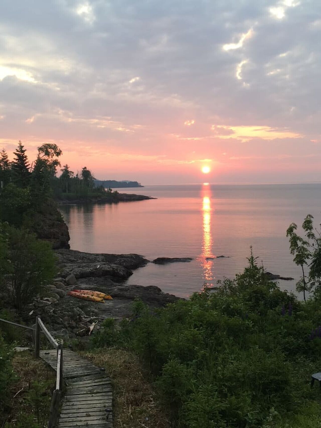 A sunset over a calm body of water with a wooden walkway leading to a rocky shoreline.