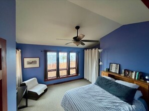 Une chambre à coucher comprenant un lit, une chaise, un bureau, un ventilateur de plafond et une fenêtre avec des rideaux.