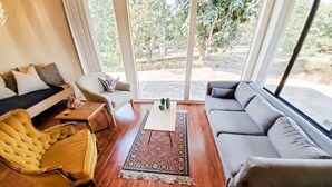 A living room with a sofa, armchair, and coffee table, large windows with a view of trees, and a wooden floor. - Guest Wing at Dairy Goat Farm (Somis)