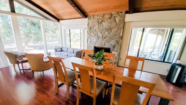 A dining area with a wooden table and chairs, a stone fireplace, and large windows. - Guest Wing at Dairy Goat Farm (Somis)
