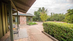 A covered outdoor area with a brick building, a paved walkway, and a neatly trimmed hedge. - Guest Wing at Dairy Goat Farm (Somis)