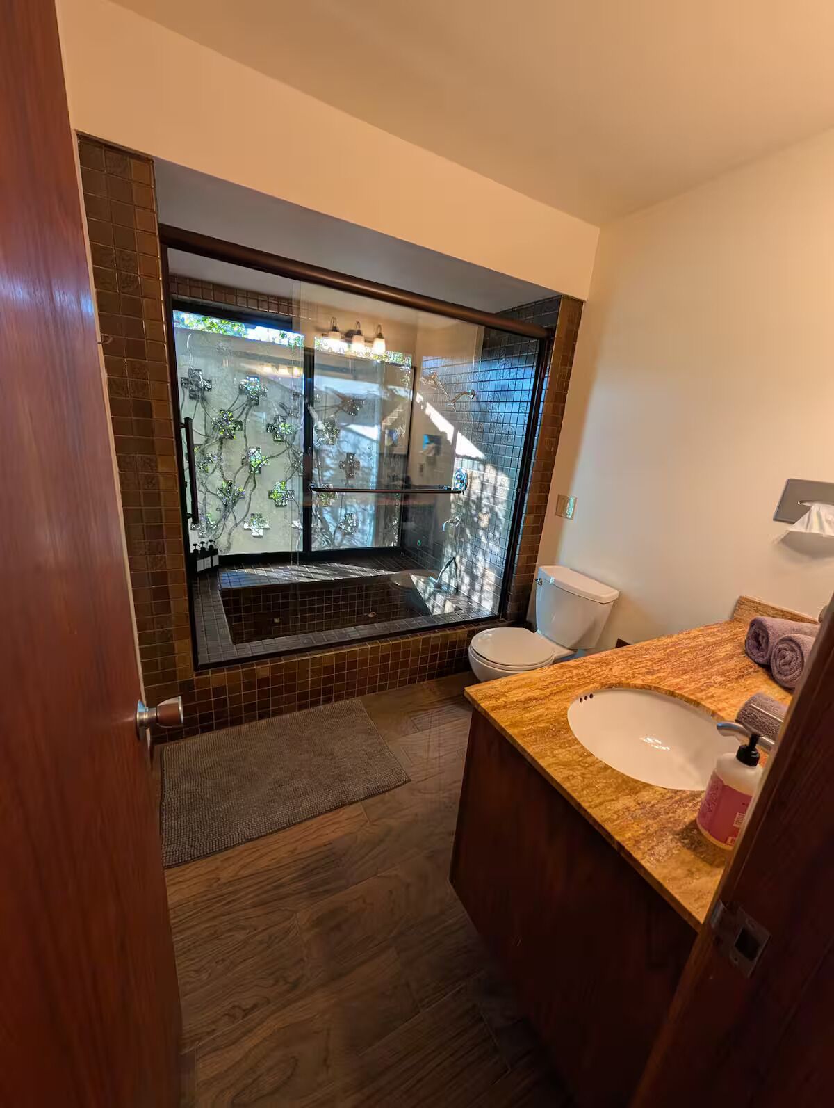 A modern bathroom with a large glass shower, a wooden vanity, and a view of a balcony.