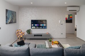 Ein modernes Wohnzimmer mit einem grauen Sofa, einem bunten Sessel, einem flachbildfernseher an der Wand und einem kleinen Tisch mit Früchten und einer Flasche.