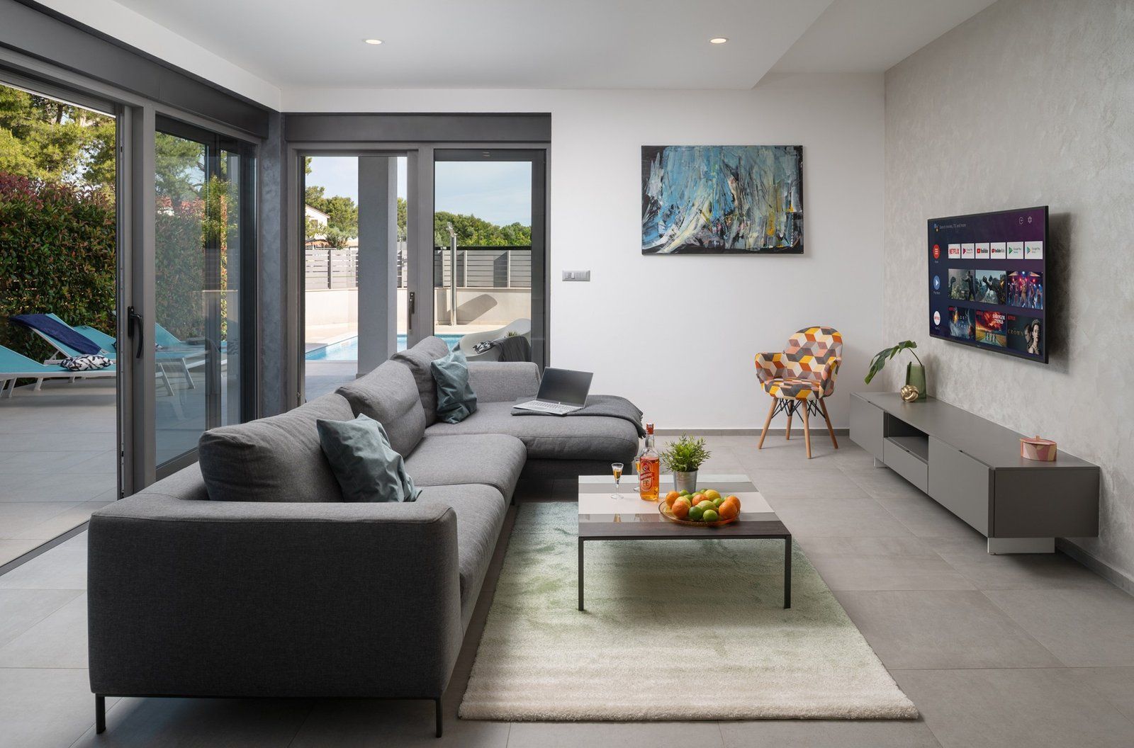 Ein modernes Wohnzimmer mit einem grauen Ecksofa, einem Couchtisch mit Obst, einem an der Wand montierten Fernseher und einer Glasschiebetür, die zu einem Poolbereich führt.