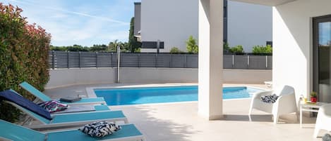 A rooftop pool area with lounge chairs and a small table, surrounded by a white building and greenery.